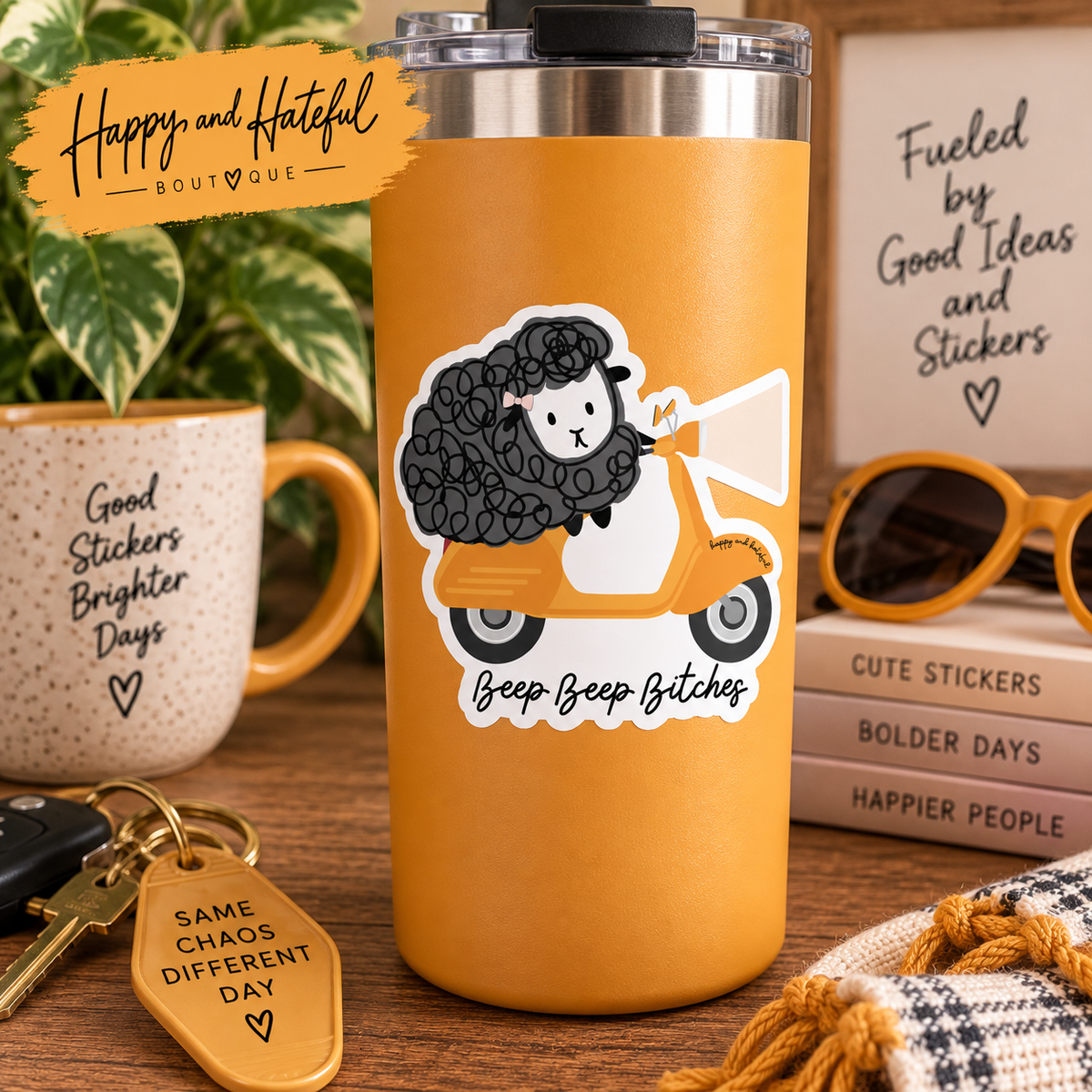 An insulated tumbler with a clear lid stands on a wooden table surrounded by lifestyle props, including a mug, sunglasses, books, and a keychain. The tumbler features a die-cut sticker of a cartoon sheep with curly gray/black hair riding an orange/yellow scooter, with the text 'Beep Beep Bitches'.