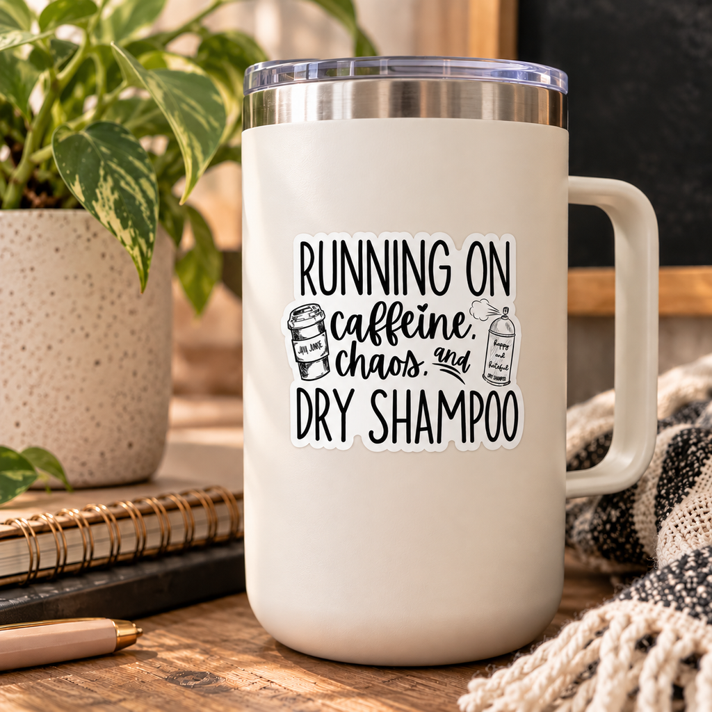 A cream-colored insulated travel mug with a clear lid sits on a wooden table surrounded by lifestyle props, including a potted plant, a notebook, and a knitted blanket. The mug features a black graphic print that reads 'RUNNING ON caffeine, chaos, and DRY SHAMPOO' with small illustrations of a coffee cup and a bottle of shampoo.