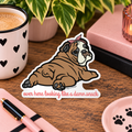 A die-cut sticker featuring a cartoon bulldog wearing a pink bow/bandana and lying down with the text 'over here looking like a damn snack' in pink script. The sticker is displayed on a wooden desk surrounded by lifestyle props including a coffee mug, notebook, and plant.