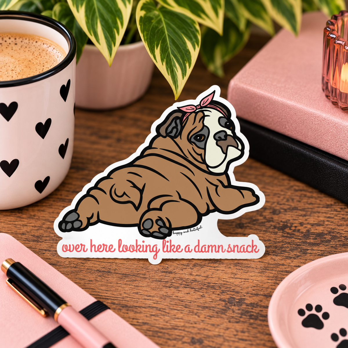 A die-cut sticker featuring a cartoon bulldog wearing a pink bow/bandana and lying down with the text 'over here looking like a damn snack' in pink script. The sticker is displayed on a wooden desk surrounded by lifestyle props including a coffee mug, notebook, and plant.