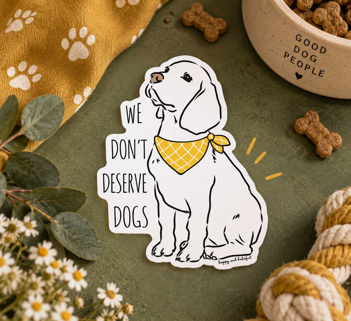 A die-cut sticker featuring a white Labrador Retriever wearing a yellow bandana, next to the text 'WE DON'T DESERVE DOGS', styled on a green surface with dog treats, a bowl, and decorative props.