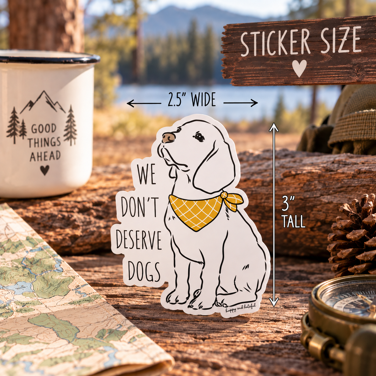 A die-cut sticker featuring a white Labrador retriever wearing a yellow bandana with the text 'WE DON'T DESERVE DOGS' is displayed outdoors on a wooden surface. The image includes dimension arrows indicating the sticker.
