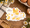 A die-cut sticker featuring a cute cartoon hen and chicks with the text 'WHAT A FUSTERCLUCK!' is displayed on a rustic wooden table surrounded by eggs, daisies, and a wooden scoop of feed.