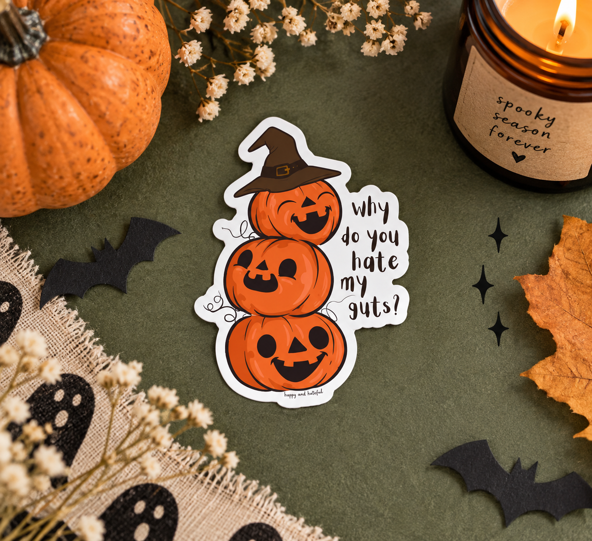 A Halloween-themed die-cut sticker featuring three stacked jack-o'-lanterns wearing a witch hat with the text 'why do you hate my guts?' on a dark olive green background. The scene is styled as a flat lay with a lit candle, a small pumpkin, dried flowers, black bat cutouts, ghost fabric, and an autumn leaf.