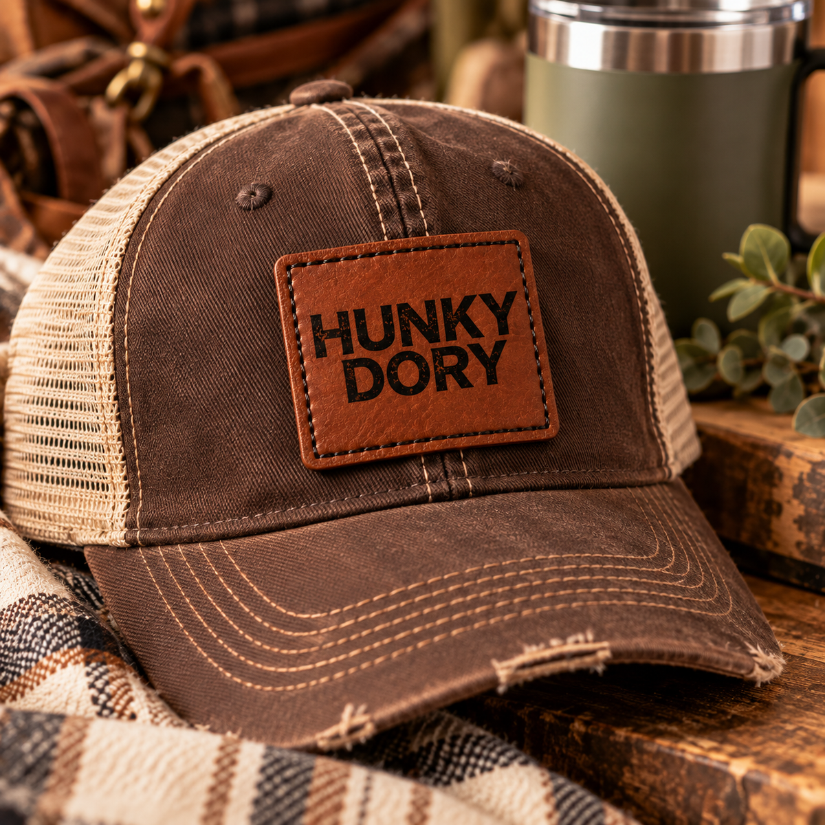 A brown distressed trucker hat with beige mesh side panels and a brown patch on the front that reads 'HUNKY DORY' in black letters. The hat is styled on a rustic wooden surface with a plaid blanket and a green travel mug in the background.