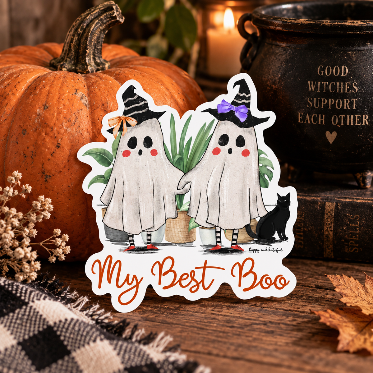 A die-cut sticker featuring two cute cartoon ghosts dressed as witches with the text 'My Best Boo' at the bottom, displayed on a rustic wooden surface with a pumpkin, books, and a cauldron in the background.