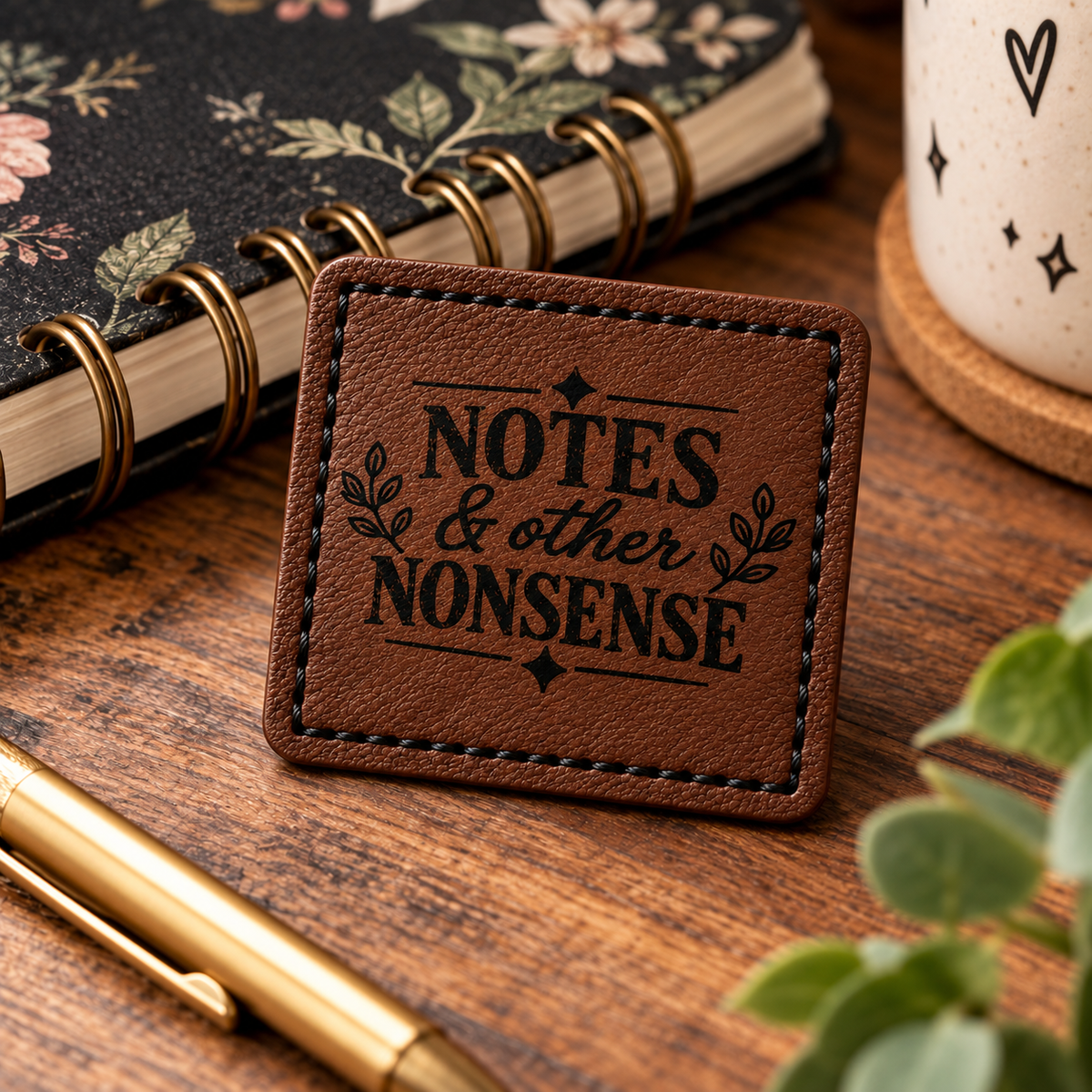 A square patch with the engraved text 'NOTES & other NONSENSE' and small leaf motifs, placed on a wooden desk with a floral notebook, pen, mug, and plant visible around it.