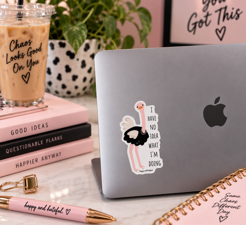 A die-cut vinyl sticker featuring a cartoon ostrich wearing a pearl necklace with  the text 'I HAVE NO IDEA WHAT I'M DOING' , shown applied to the back of a silver laptop. The laptop is styled on a marble desk with a pink iced coffee, books, a pen, and a notebook.