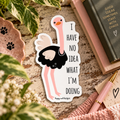 A die-cut sticker featuring a cartoon ostrich wearing a pearl necklace with  the text 'I HAVE NO IDEA WHAT I'M DOING' on a white background, styled on a rustic wooden surface with a notebook, pen, and green leaves.