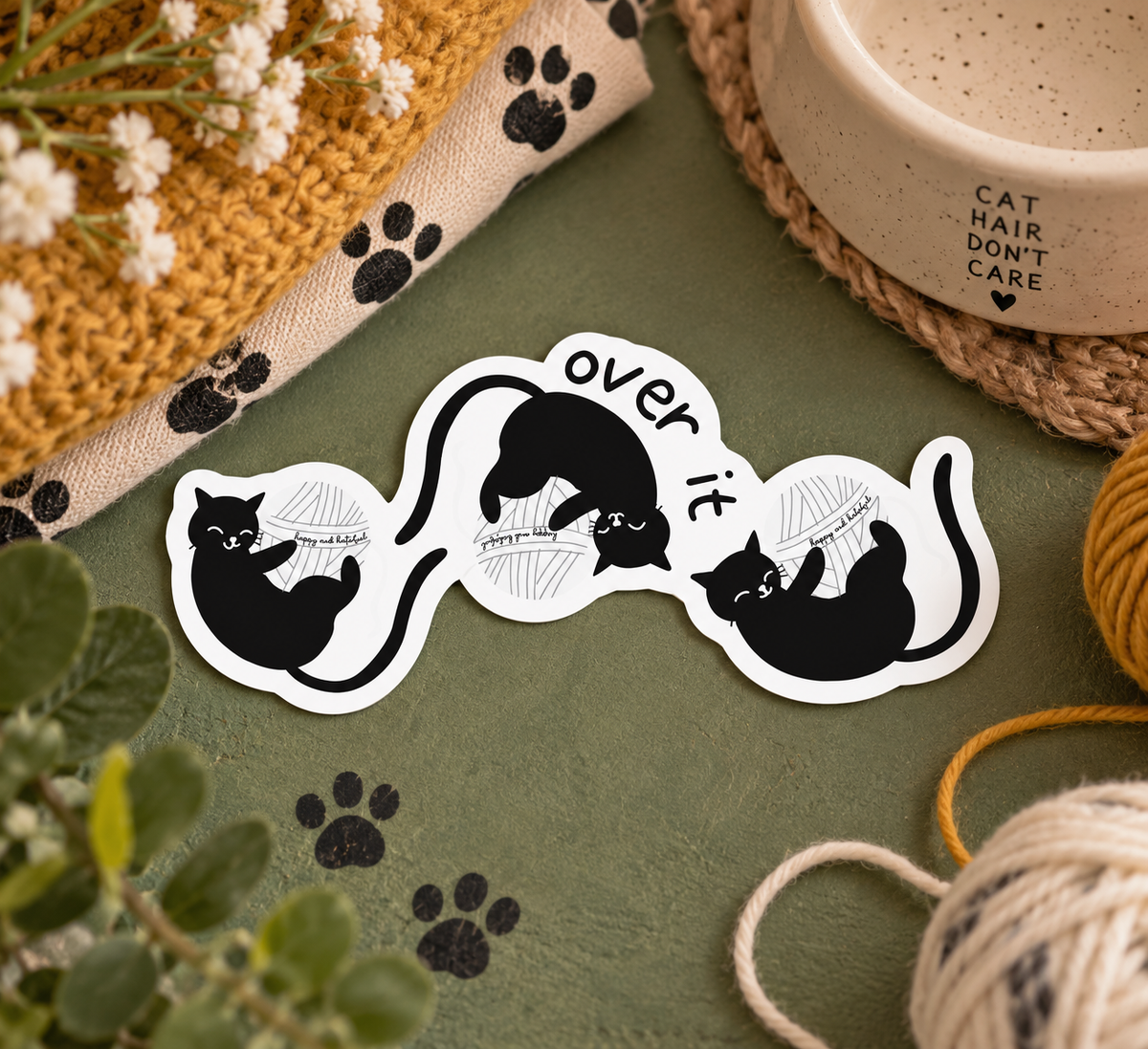 A die-cut vinyl sticker featuring black cartoon cats playing with yarn and the text 'over it', displayed on a textured olive green surface with cat-themed props including yarn balls, a ceramic mug, and a woven hat.