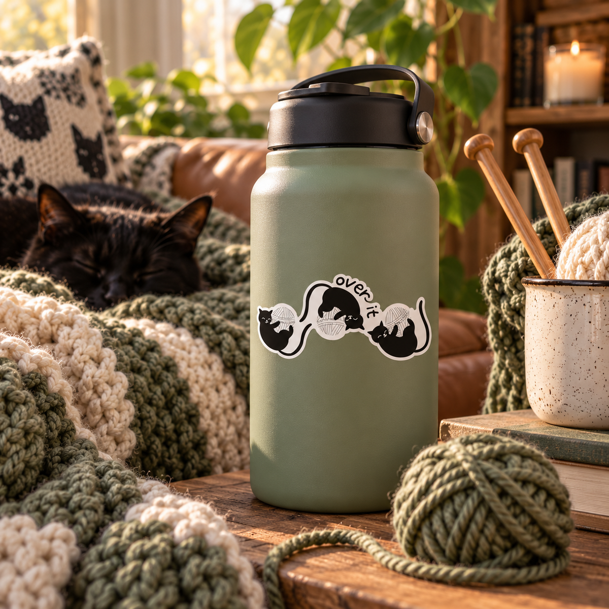 A matte sage green insulated water bottle with a black lid and sticker featuring three black cartoon cats playing with white yarn balls, with the text 'over it' stands on a wooden table in a cozy living room. A black cat rests on a chunky knit blanket in the background, and a yarn ball, knitting needles, and a mug are nearby.