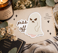 A die-cut sticker featuring a ghost with an angry expression and the words 'piss off' in brown script, placed on a styled flat-lay with a candle, dried flowers, eucalyptus leaves, a notebook, and a plaid fabric.