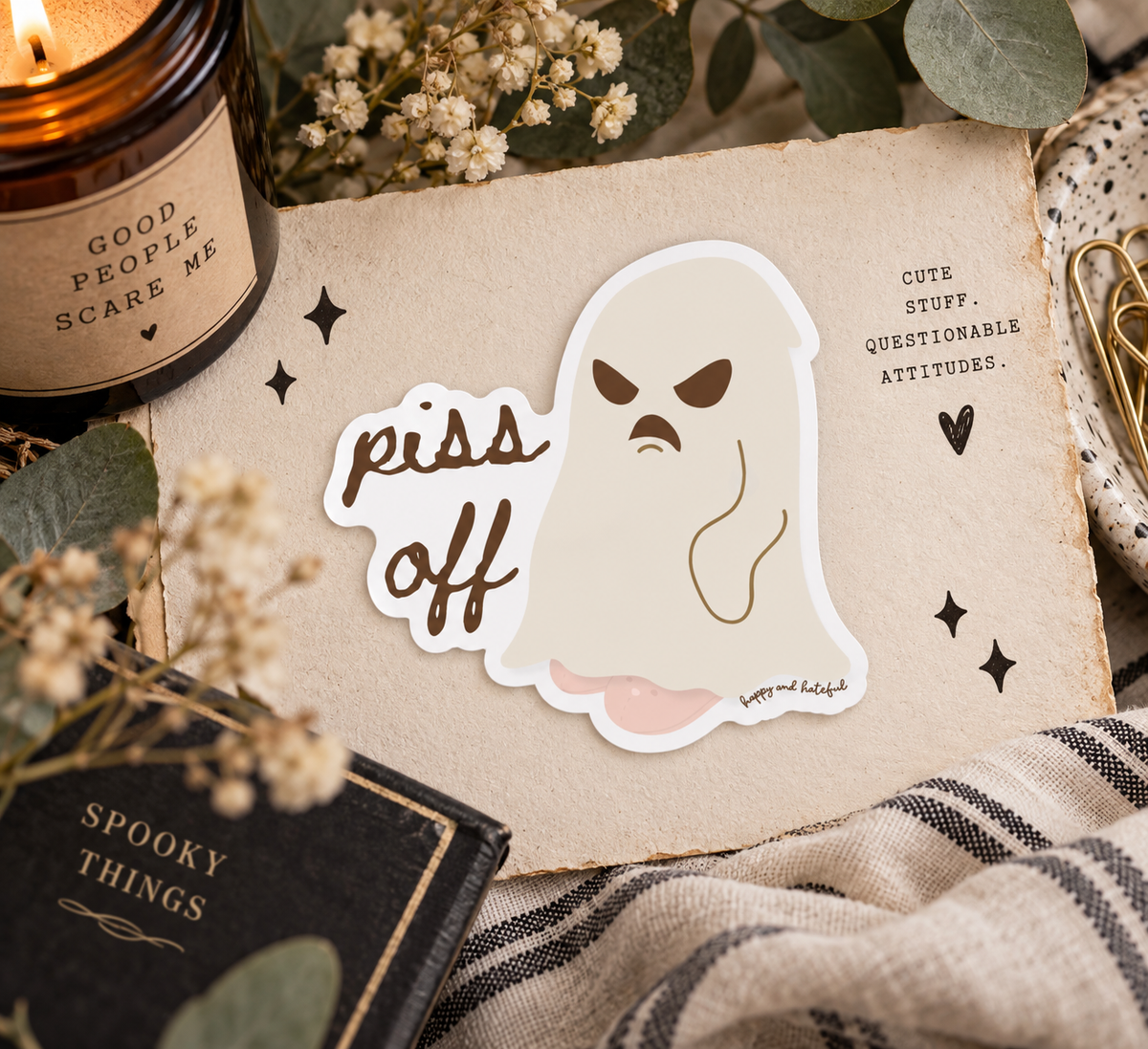 A die-cut sticker featuring a ghost with an angry expression and the words 'piss off' in brown script, placed on a styled flat-lay with a candle, dried flowers, eucalyptus leaves, a notebook, and a plaid fabric.