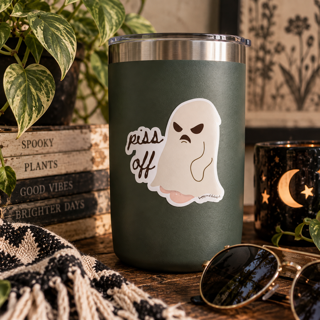 An insulated tumbler cup with a dark green body, stainless steel rim, and a die-cut sticker of a ghost with an angry expression and the words 'piss off' on the front. The cup is styled on a wooden table with a knit blanket, books, a plant, and sunglasses in a cozy autumn setting.