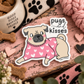 A die-cut sticker featuring a cute cartoon pug wearing a pink sweater with white hearts and the words 'pugs and kisses' above it, displayed on a rustic pink wooden surface with dog treats, a dog bowl, and pink yarn around it.