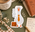 A die-cut sticker featuring a cartoon goose wearing oversized round glasses, a tie, and carrying a briefcase, with the text 'TODAY I'M A SERIOUS GOOSE'. The sticker is displayed in a styled flat-lay setting on a dark green surface, surrounded by a ceramic mug, a brown book, a checkered cloth, and a notepad.