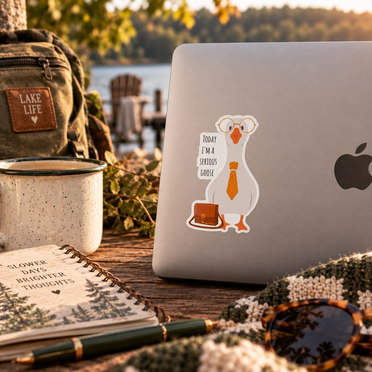 A silver laptop with a cartoon goose wearing oversized round glasses, a tie, and carrying a briefcase, with the text 'TODAY I'M A SERIOUS GOOSE' is set on a wooden table outdoors near water. Surrounding props include a green backpack, a speckled mug, an open notebook, a pen, sunglasses, and a knit blanket.