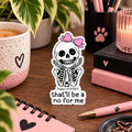A die-cut sticker featuring a skeleton with a pink bow adorning a skull and the words 'That'll be a no for me' below it, styled on a rustic wooden surface with in a cozy pink, black, and botanical desk setting.