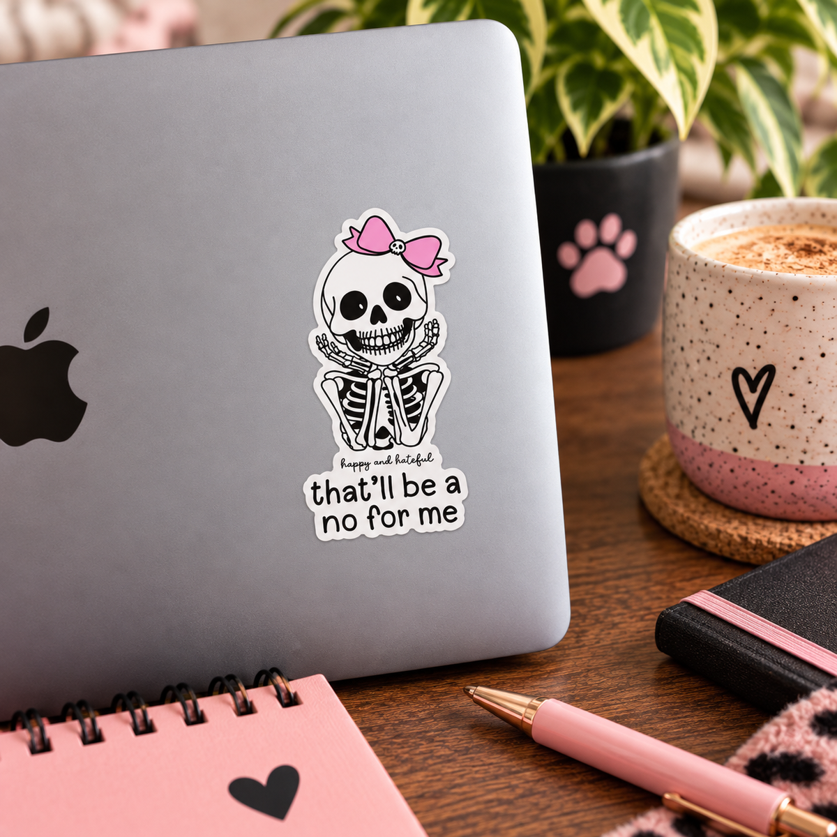A die-cut sticker featuring a skeleton with a pink bow adorning a skull and the words 'That'll be a no for me' below it placed on a lap top, styled on a rustic wooden surface with in a cozy pink, black, and botanical desk setting.