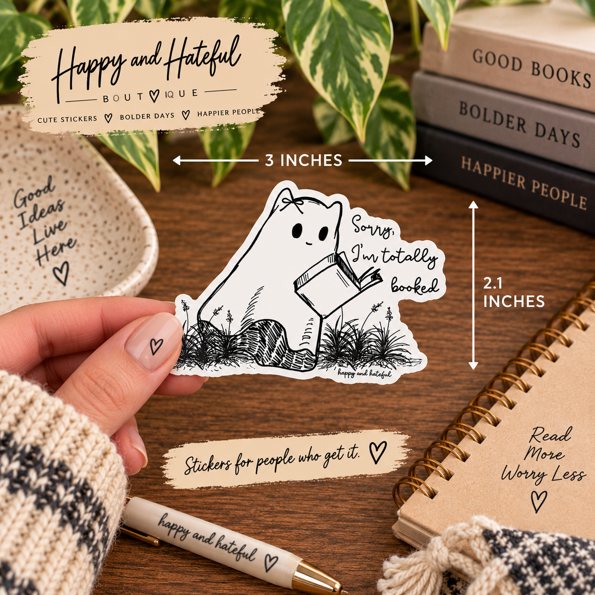 A hand holds a die-cut sticker featuring a cute black-and-white cartoon ghost cat reading a book with the text "Sorry, I'm totally booked." The scene is styled on a wooden desk with notebooks, books, and decorative text overlays showing the sticker dimensions as 3 inches by 2.