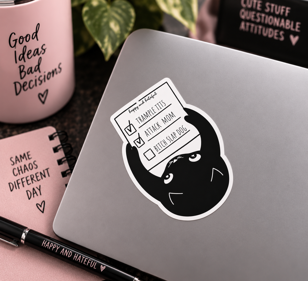 A die-cut sticker featuring a black cartoon cat holding a checklist with items 'TRAMPLE TIT'S', 'ATTACK MOM' both check marked done and 'BITCH SLAP DOG' unchecked, shown applied to the back of a silver laptop. The scene is styled with a pink mug, notebook, plant, and pen on a dark tabletop.