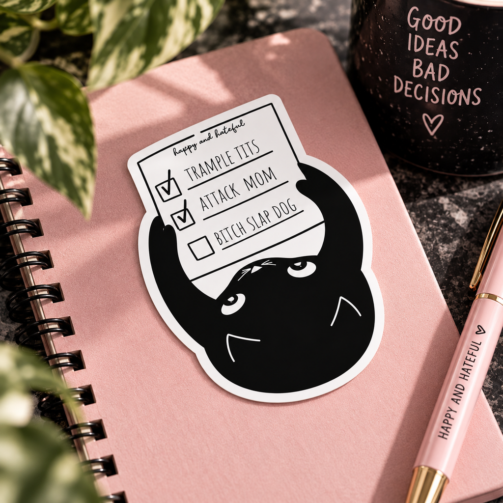 A die-cut sticker featuring a black cartoon cat holding a checklist with items 'TRAMPLE TIT'S', 'ATTACK MOM' both check marked done and 'BITCH SLAP DOG' unchecked, placed on a pink notebook with decorative plants, a black mug, and a pink pen.