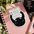 A die-cut sticker featuring a black cartoon cat holding a checklist with items 'TRAMPLE TIT'S', 'ATTACK MOM' both check marked done and 'BITCH SLAP DOG' unchecked, placed on a pink notebook with decorative plants, a black mug, and a pink pen.