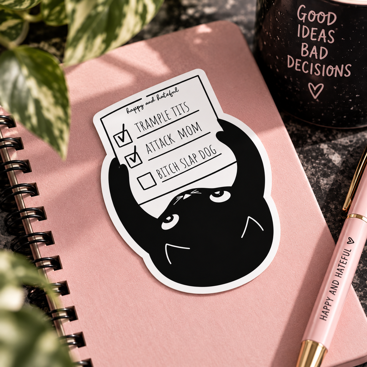 A die-cut sticker featuring a black cartoon cat holding a checklist with items 'TRAMPLE TIT'S', 'ATTACK MOM' both check marked done and 'BITCH SLAP DOG' unchecked, placed on a pink notebook with decorative plants, a black mug, and a pink pen.