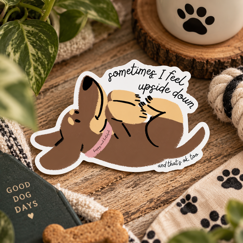 A die-cut sticker featuring a cartoon dog lying upside down with the text 'sometimes I feel upside down and that's ok, too', placed on a wooden surface with dog-themed props including a book, rope, and paw-print fabric.