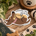 A die-cut sticker featuring a cartoon dog lying upside down with the text 'sometimes I feel upside down and that's ok, too', placed on a wooden surface with dog-themed props including a book, rope, and paw-print fabric.