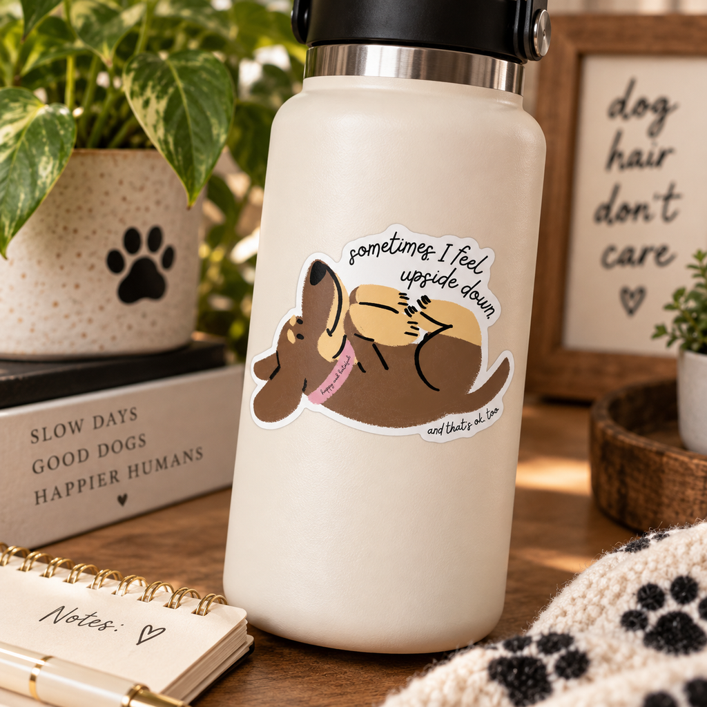A cream-colored insulated water bottle with a black lid, featuring a cartoon dog sticker that reads 'sometimes I feel upside down and that's ok too'. The bottle is styled on a wooden table with a notebook, a plant, and a cozy background.