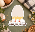 A die-cut sticker featuring a cartoon eggshell cracking open to reveal the bottom half of a chicken, with the text 'WHAT THE SHELL?'.  The sticker is displayed on a textured green surface surrounded by eggs, a broken eggshell, daisies, and a plaid cloth.