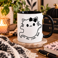 A white ceramic mug with a black interior and handle, featuring a cute black line-art cat ghost design with the text 'you do you, boo' wrapped around it. The mug sits on a wooden coaster on a rustic table surrounded by cozy lifestyle props including a lit candle, a knit blanket, books, and a notebook.