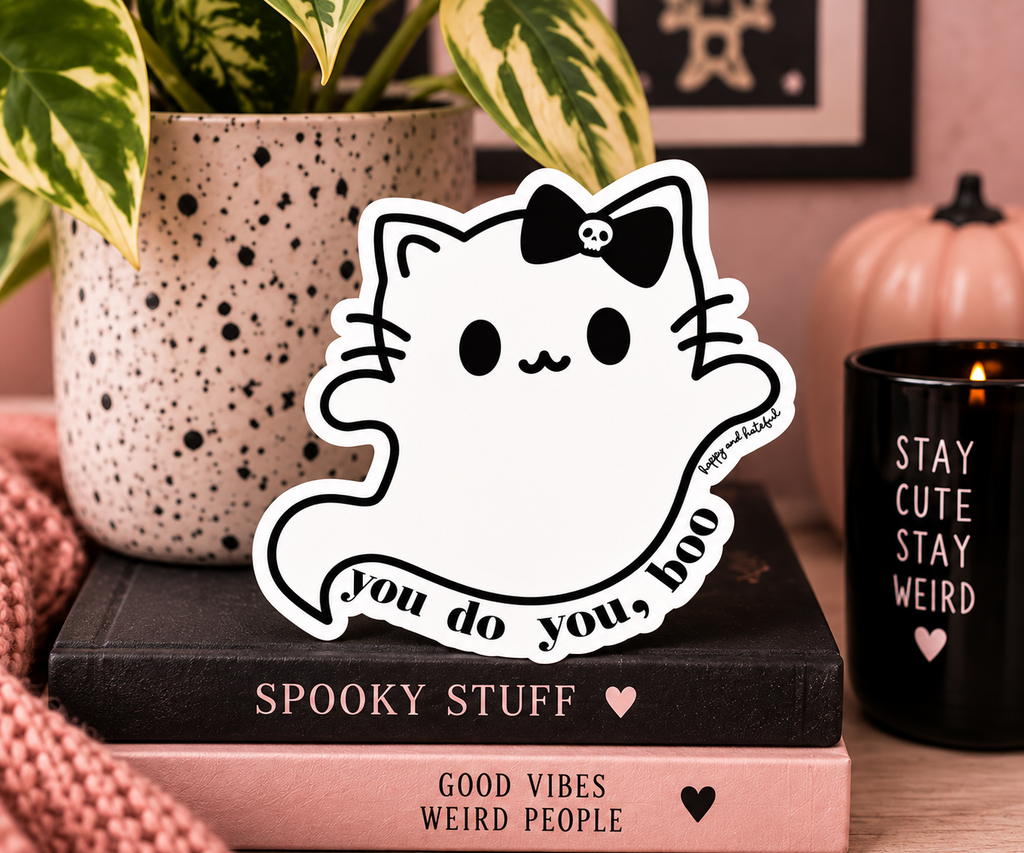 A die-cut vinyl sticker featuring a cute white cartoon ghost cat with a skull-adorned bow and the text 'You do you, boo', displayed on a stack of books in a cozy, styled tabletop setting.