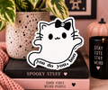 A die-cut vinyl sticker featuring a cute white cartoon ghost cat with a skull-adorned bow and the text 'You do you, boo', displayed on a stack of books in a cozy, styled tabletop setting.