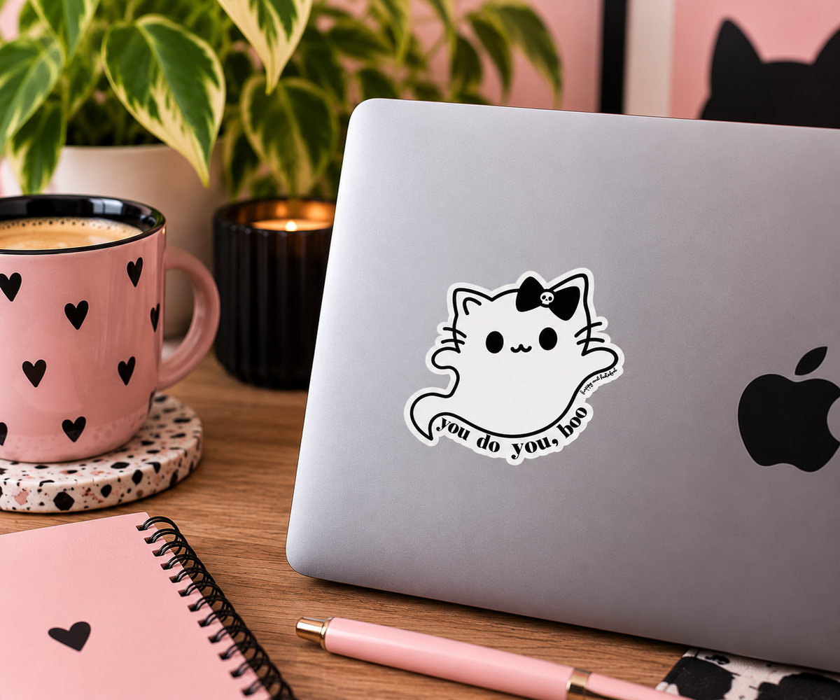 A white cartoon ghost cat sticker with a skull-adorned black bow and the text 'you do you, too' applied to the back of a silver laptop. The laptop sits on a wooden desk in a cozy, styled workspace setting with a pink heart-patterned mug, a notebook, and a plant in the background.