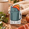 A die-cut sticker featuring a cartoon blue-gray cat wearing an orange and yellow beanie with the text 'YOU'RE LAME' in script. The scene is styled as a cozy wooden table, white and orange speckled mug, a plant, and an cream knit blanket.