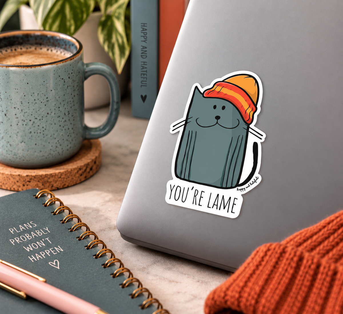 A die-cut sticker featuring a cartoon blue-gray cat wearing an orange and yellow beanie with the text 'YOU'RE LAME' in script, shown applied to the back of a gray laptop. The scene is styled as a cozy desk flat-lay with a blue speckled mug, a notebook, and an orange knit sweater.